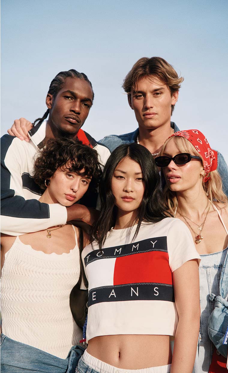 Group of five young adults posing together wearing Tommy Jeans, including crop tops and jackets, against a clear sky background.