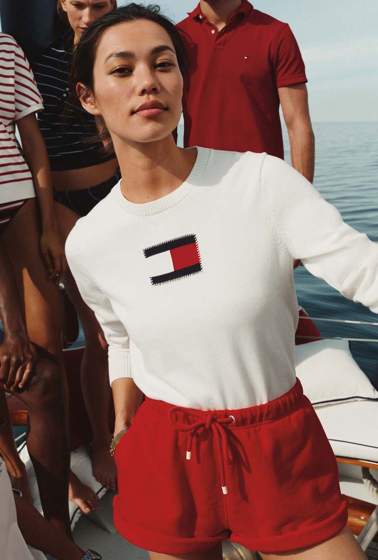 Woman wearing a white Tommy Hilfiger top and red shorts standing on a boat deck, with water in the background.