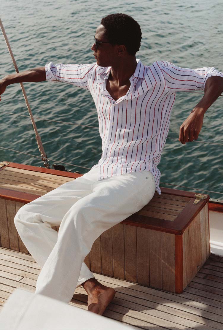 Man sitting on a wooden bench on a boat wearing a Tommy Hilfiger striped shirt and light pants, looking out over the water.