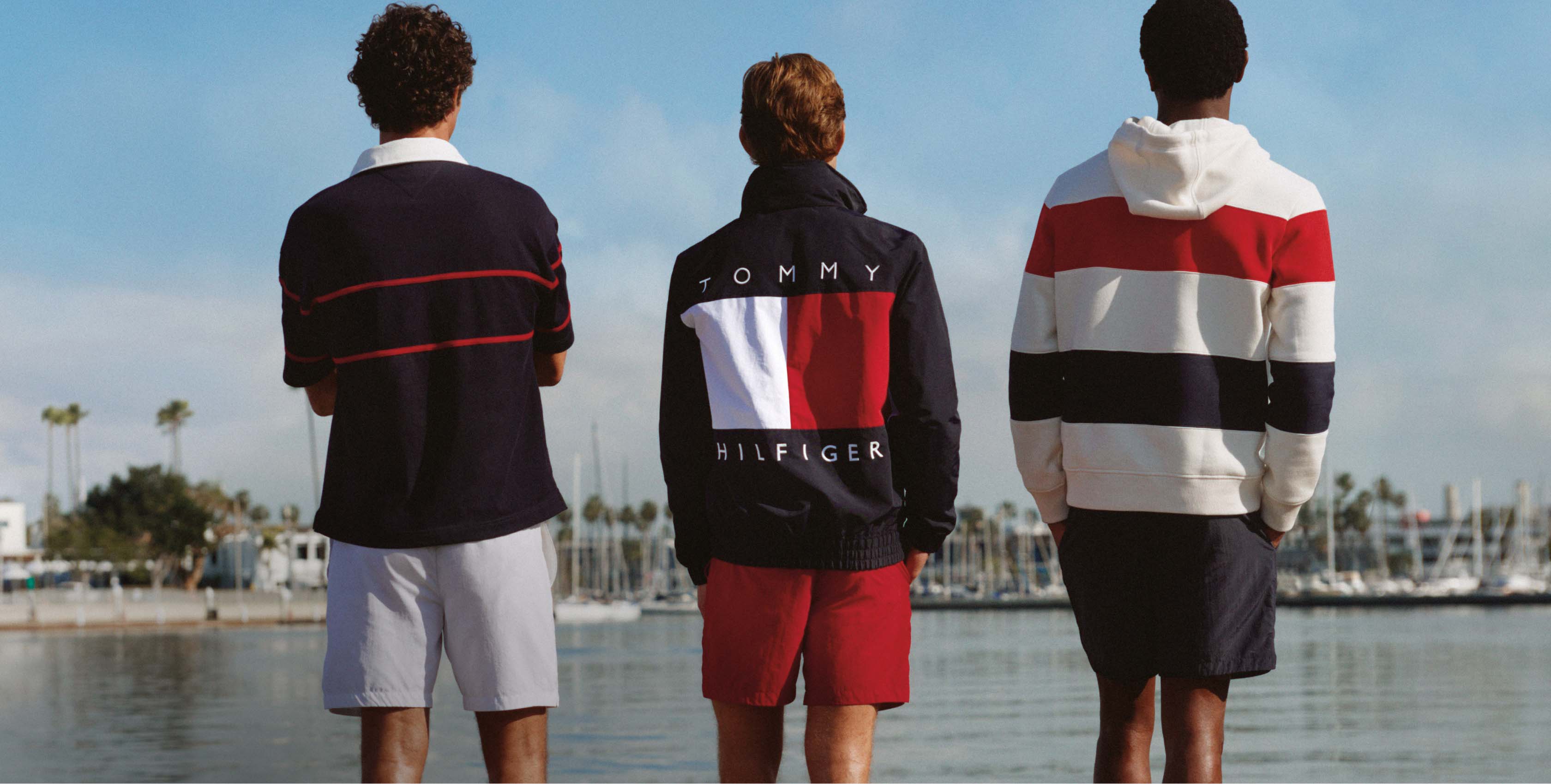 Three men standing with their backs to the camera by a marina, wearing color-blocked and striped Tommy Hilfiger outfits in red, white, and blue tones.