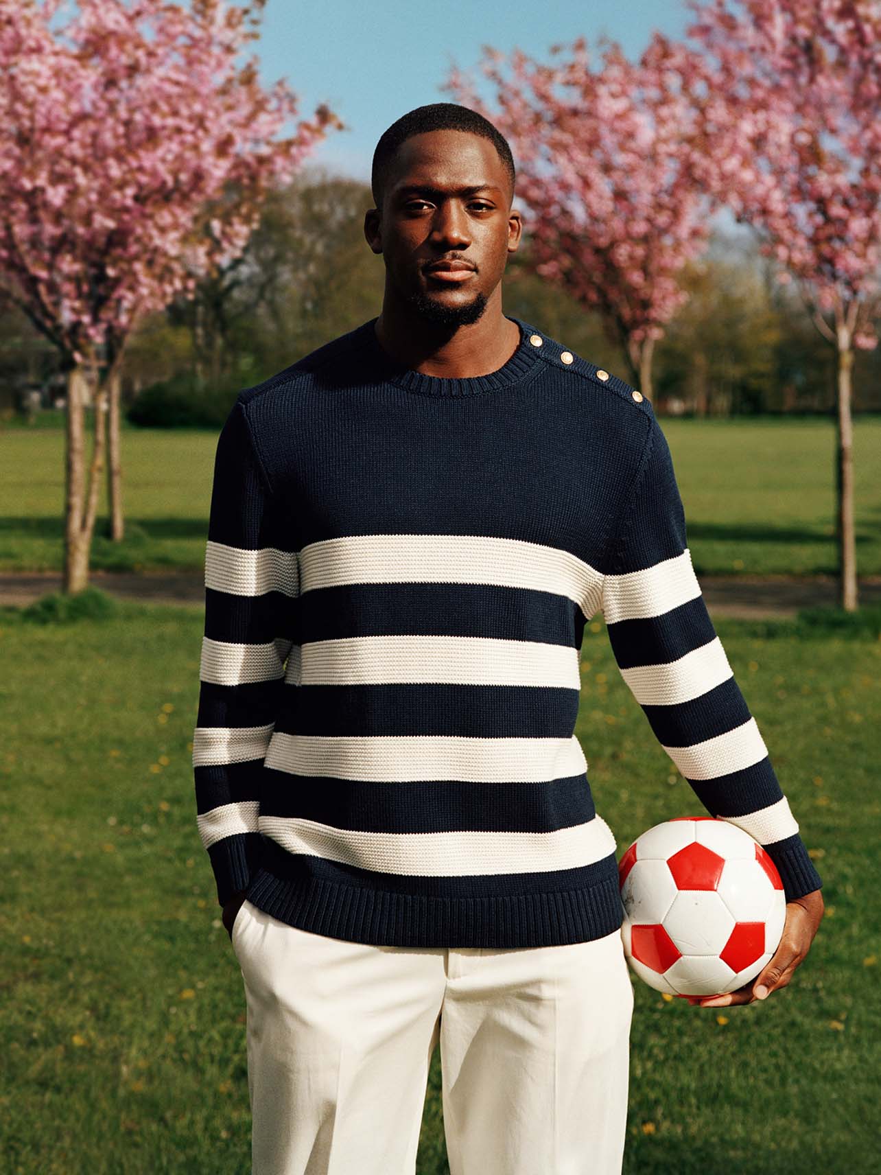 Ibrahima Konate in a park wearing a navy sweater with bold white stripes and light trousers, also holding a football.