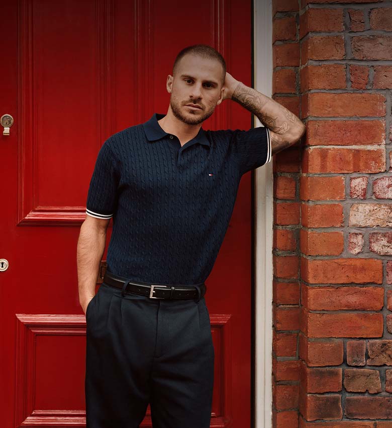 Left side Liverpool player Alexis Mcallister stand against bold red and brick backdrops wearing navy Tommy Hilfiger summer knitwear. Right side Liverpool player Cody Gakpo standing un red background wearing Tommy Hilfiger summer knitwear. 