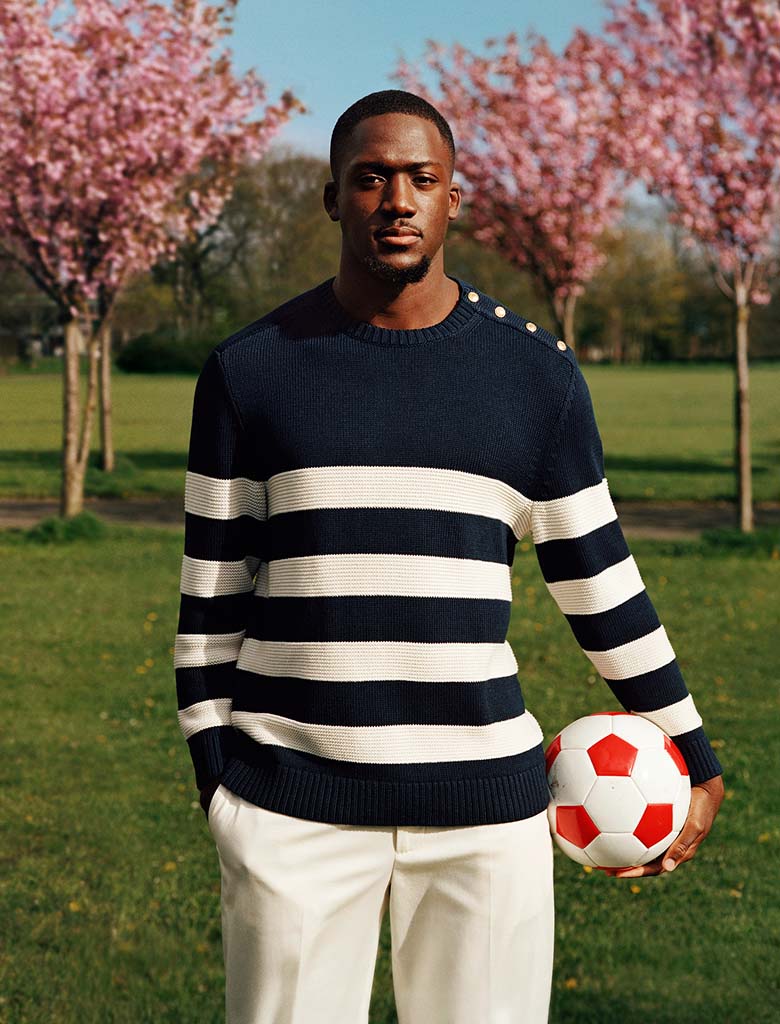 Liverpool player Ibrahima Konate standing outdoors in a park holding a football, wearing a Tommy Hilfiger navy sweater with wide white horizontal stripes and light trousers.