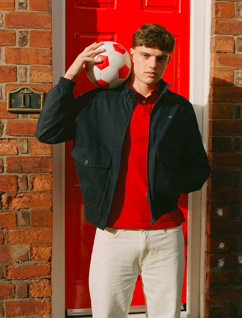 Liverpool player Giovanni Leoni  posing outside a brick house with a football on his shoulder, wearing a Tommy Hilfiger navy jacket over a red polo shirt and light trousers.