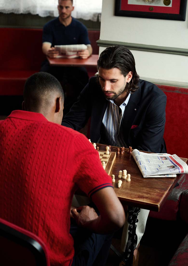 Liverpool player Dominik Szoboszlai and Alexander Isak sitting at a café table playing chess in a Liverpool FC-inspired lifestyle setting. One wears a Tommy Hilfiger red knit polo, the other a Tommy Hilfiger navy blazer.