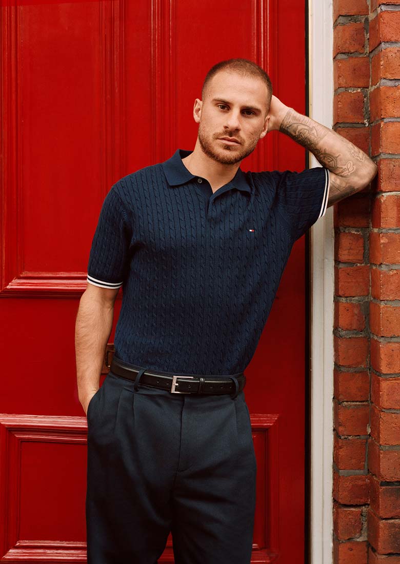 Liverpool player Alexis Mac Allister leaning against a red doorway wearing a Tommy Hilfiger navy textured polo shirt and tailored trousers. Text reads ‘Rooted in heritage, shaped by individuality.