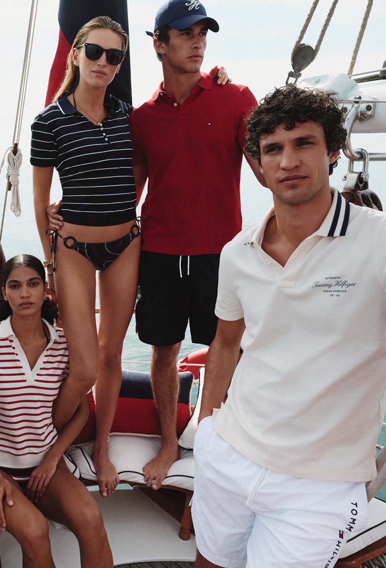 Group of six models on a sailboat wearing classic Tommy Hilfiger polo shirts and nautical-inspired outfits, posing together under a clear sky.