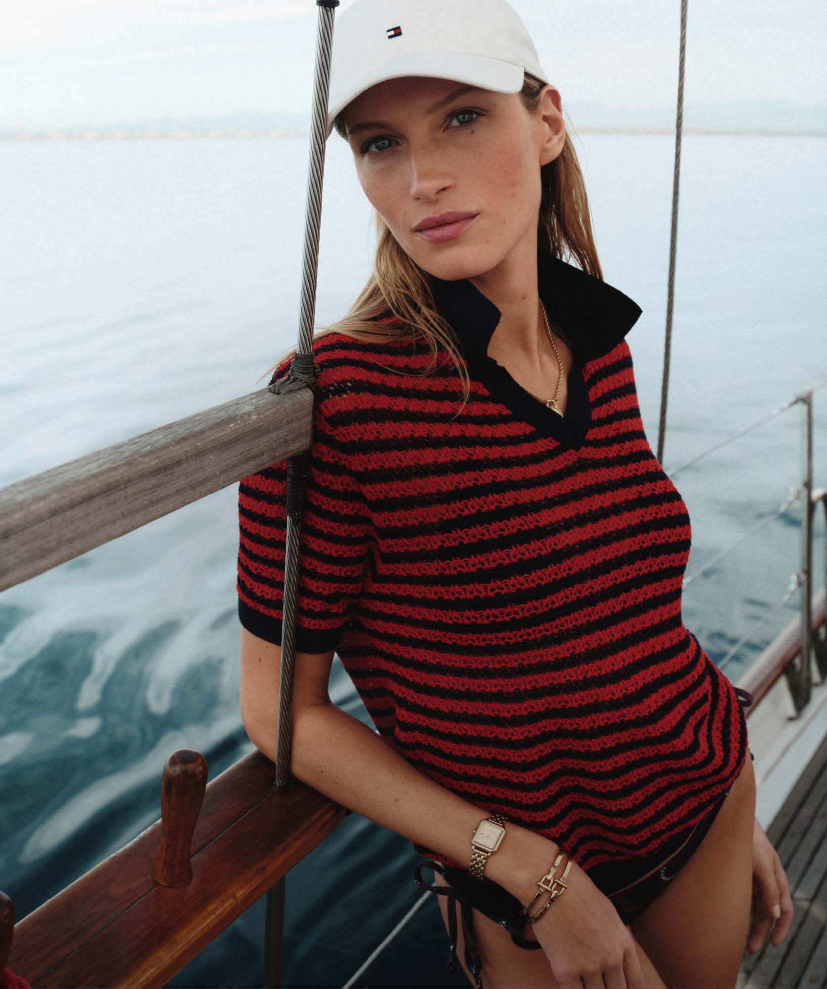 Woman leaning on a boat railing wearing a Tommy Hilfiger red and navy striped top and a white cap, with the ocean behind her.
