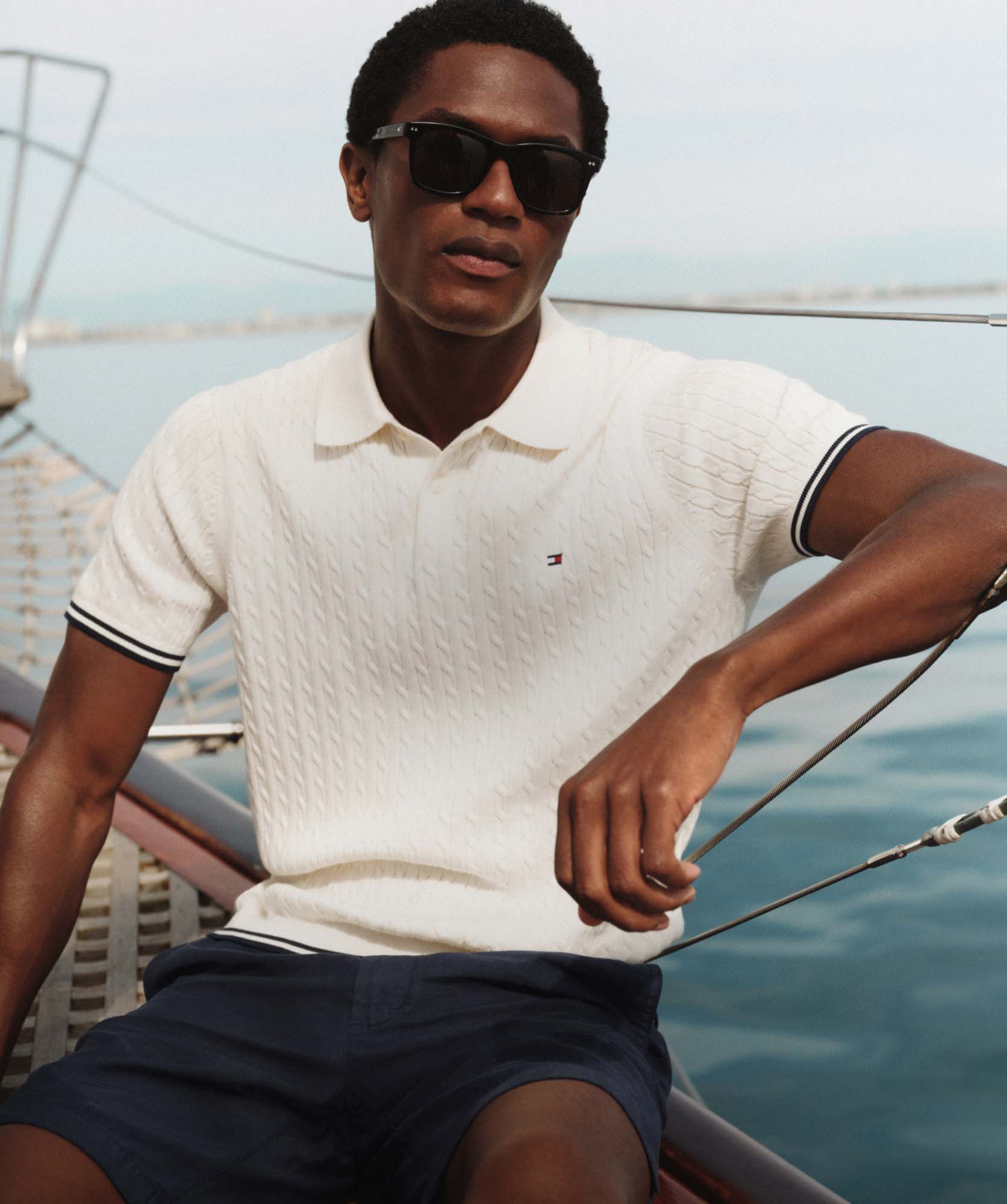 Man sitting on a boat wearing a textured Tommy Hilfiger white polo shirt, sunglasses, and navy shorts, looking relaxed by the water.