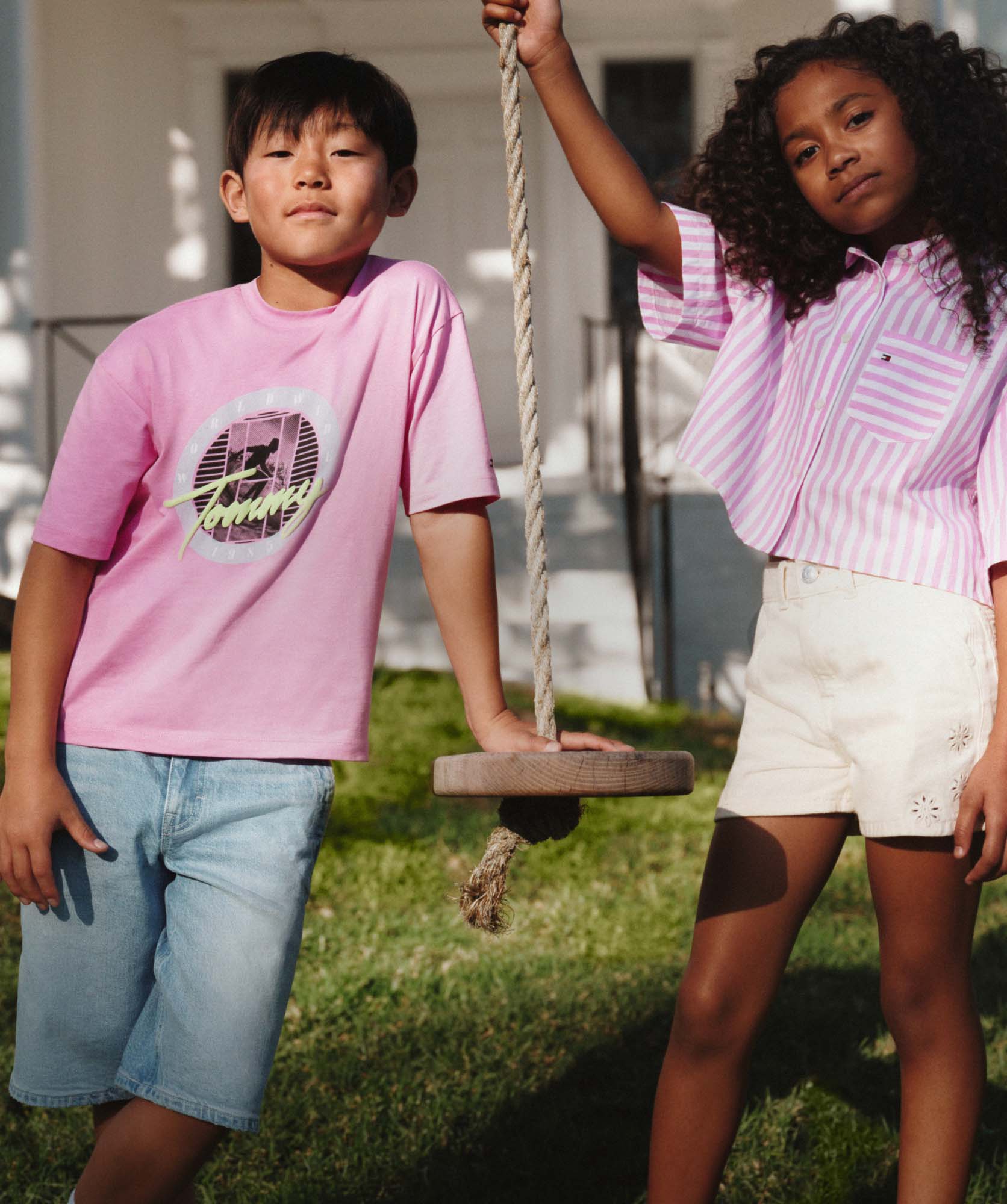 Two children standing outdoors near a rope swing, wearing casual summer Tommy Hilfiger clothes, with a house in the background.