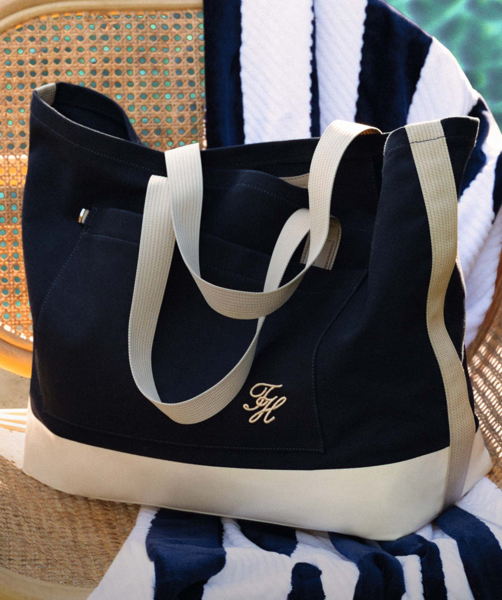 Close-up of a Tommy Hilfiger navy tote bag with cream handles placed on a striped towel, with a woven chair in the background.
