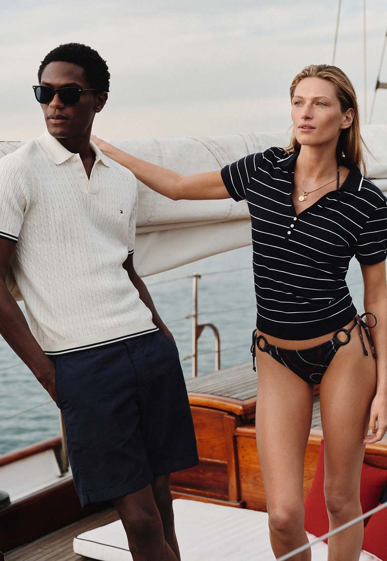 Man and woman relaxing on a sailboat deck wearing Tommy Hilfiger polo shirts, close together with the ocean in the background.