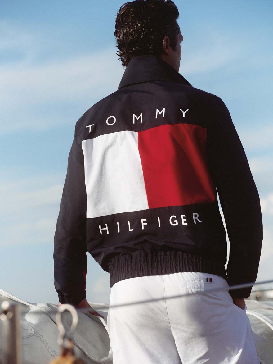Man standing on a boat facing away, wearing a Tommy Hilfiger navy jacket with a large red, white, and white Tommy Hilfiger logo panel on the back.
