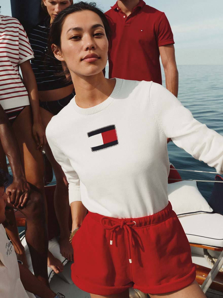 Woman standing on a boat wearing a white T-shirt with a Tommy Hilfiger logo and high-waisted red shorts, with other people and the ocean in the background.
