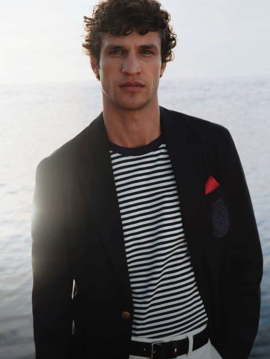 Man standing by the water wearing a Tommy Hilfiger navy blazer over a striped shirt with a red pocket square, looking at the camera.