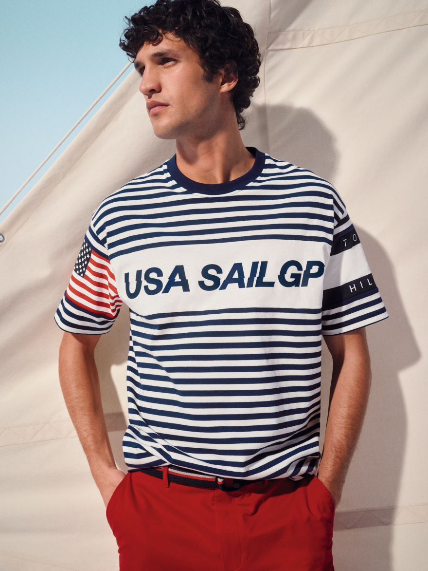 Model wearing a Tommy Hilfiger navy-and-white striped USA SailGP T-shirt paired with red shorts, standing against a sail-inspired backdrop.