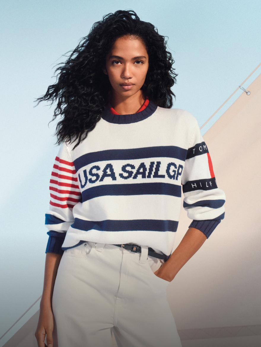 Woman model wearing a striped tommy Hilfiger USA SailGP sweater with white pants, standing outdoors beside sail canvas structures.