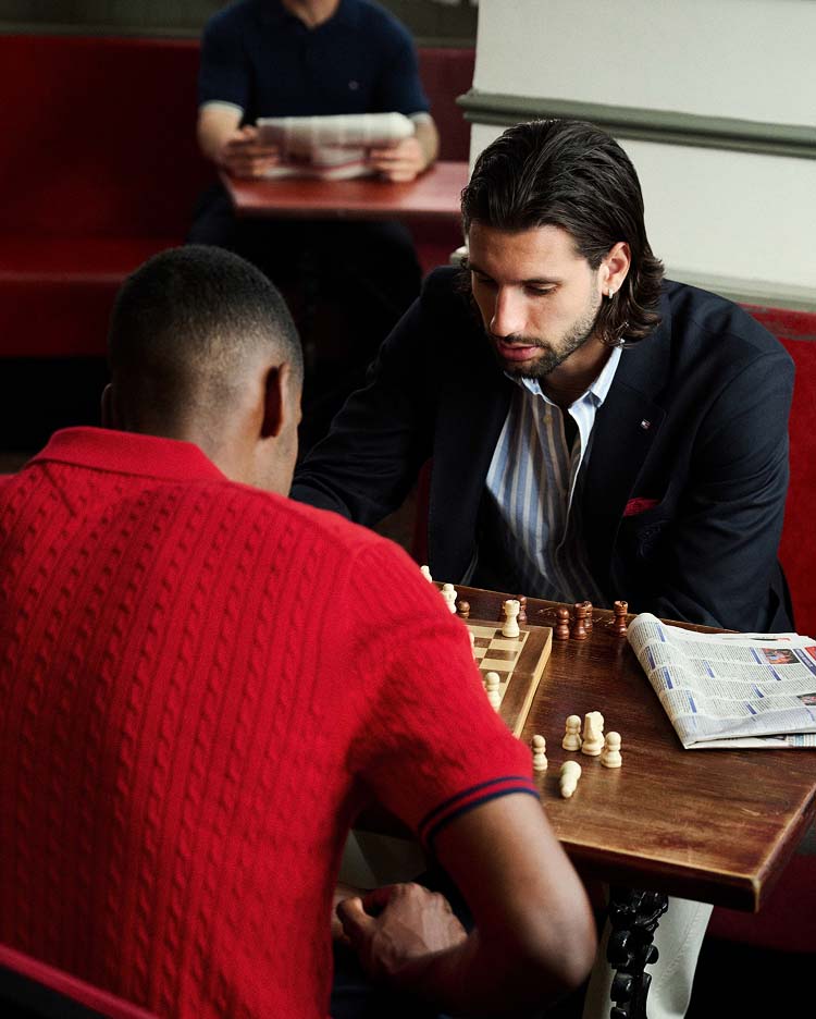 Liverpool player Dominik Szoboszlai and Alexander Isak sitting at a café table playing chess in a Liverpool FC-inspired lifestyle setting. One wears a Tommy Hilfiger red knit polo, the other a Tommy Hilfiger navy blazer.