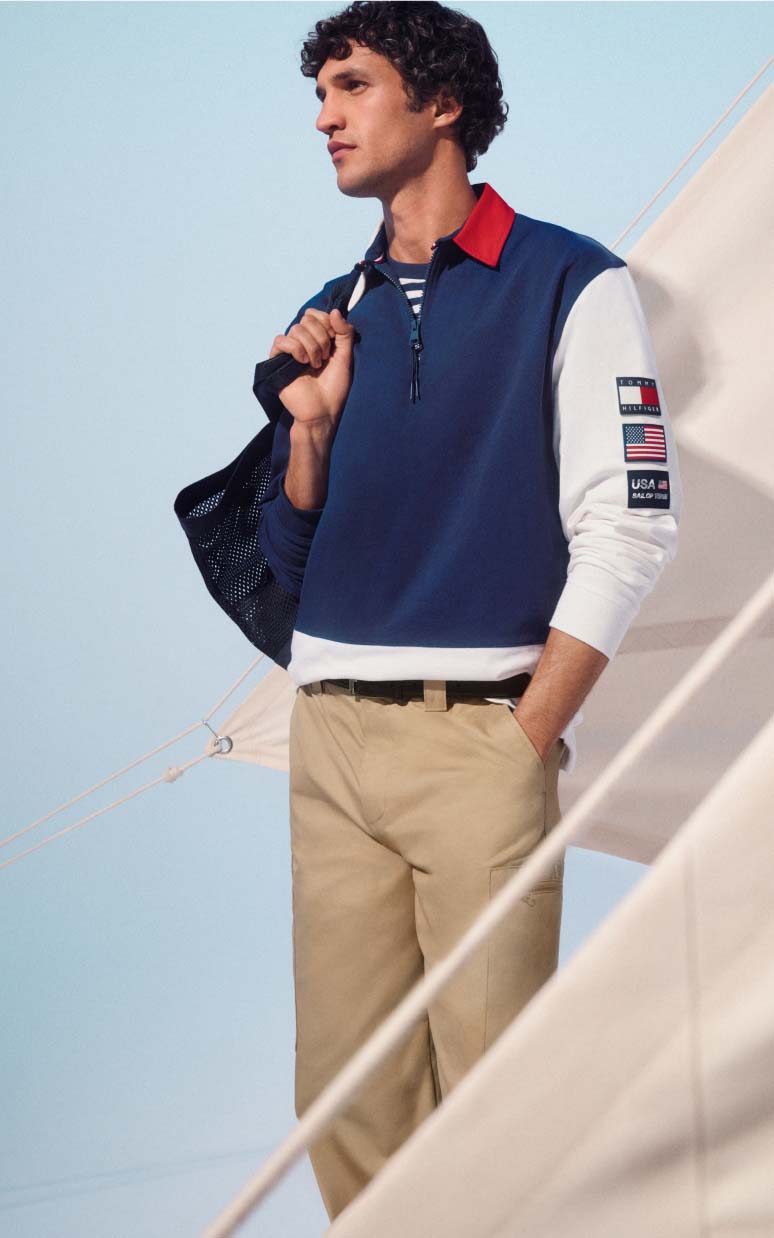 Male model wearing a Tommy x U.S. SailGP navy rugby-style shirt and khaki pants on a boat.