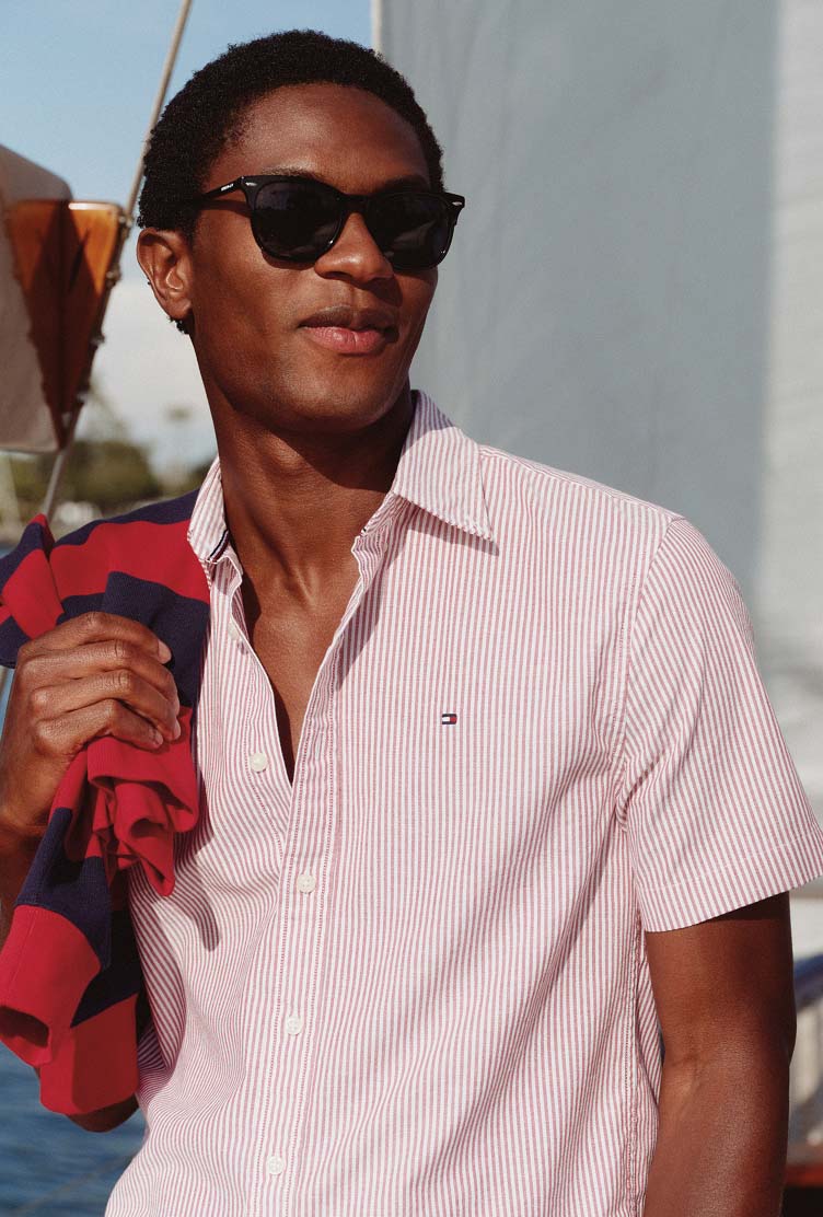 Male model in a Tommy Hilfiger pink striped button-down shirt and sunglasses standing on a boat.