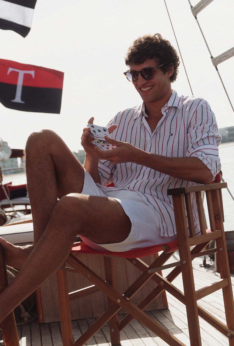 Male model seated on a wooden chair wearing a Tommy Hilfiger striped shirt and shorts on a yacht deck.