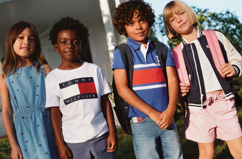Four children wearing colorful Tommy Hilfiger outfits and backpacks standing outdoors in warm sunlight.
