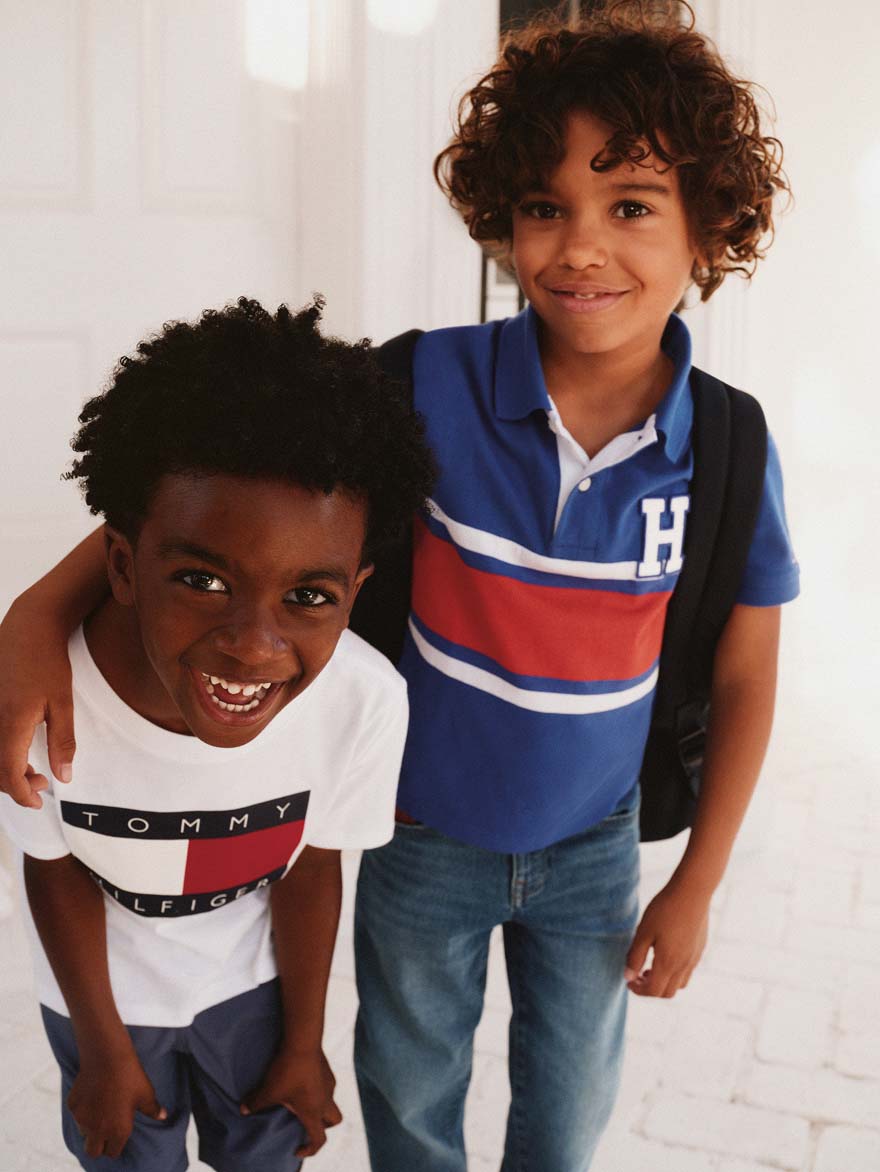Two boys smiling indoors, one wearing a white Tommy Hilfiger logo T-shirt and the other wearing a blue striped polo shirt with a backpack.