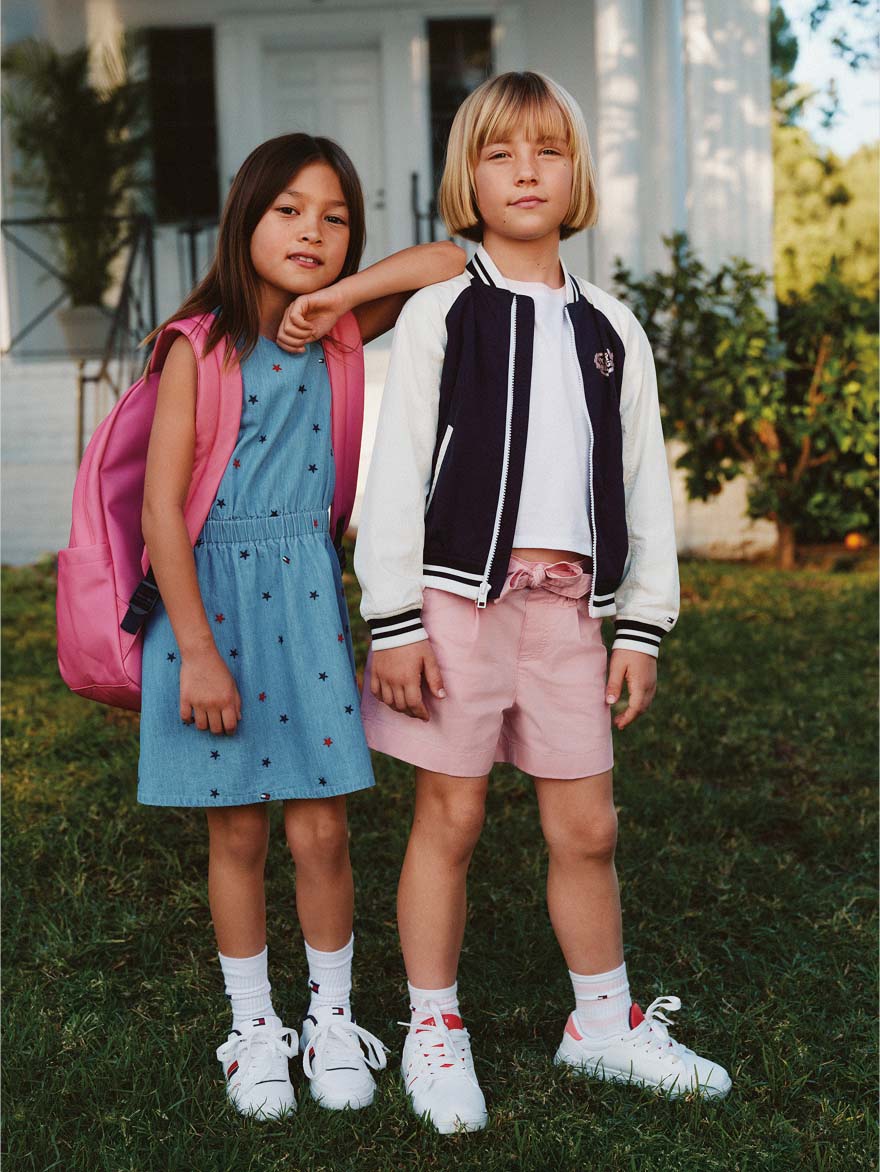 Two girls standing outside wearing coordinated pastel Tommy Hilfiger outfits, including a blue sleeveless dress and a varsity-style jacket with pink shorts and backpacks.