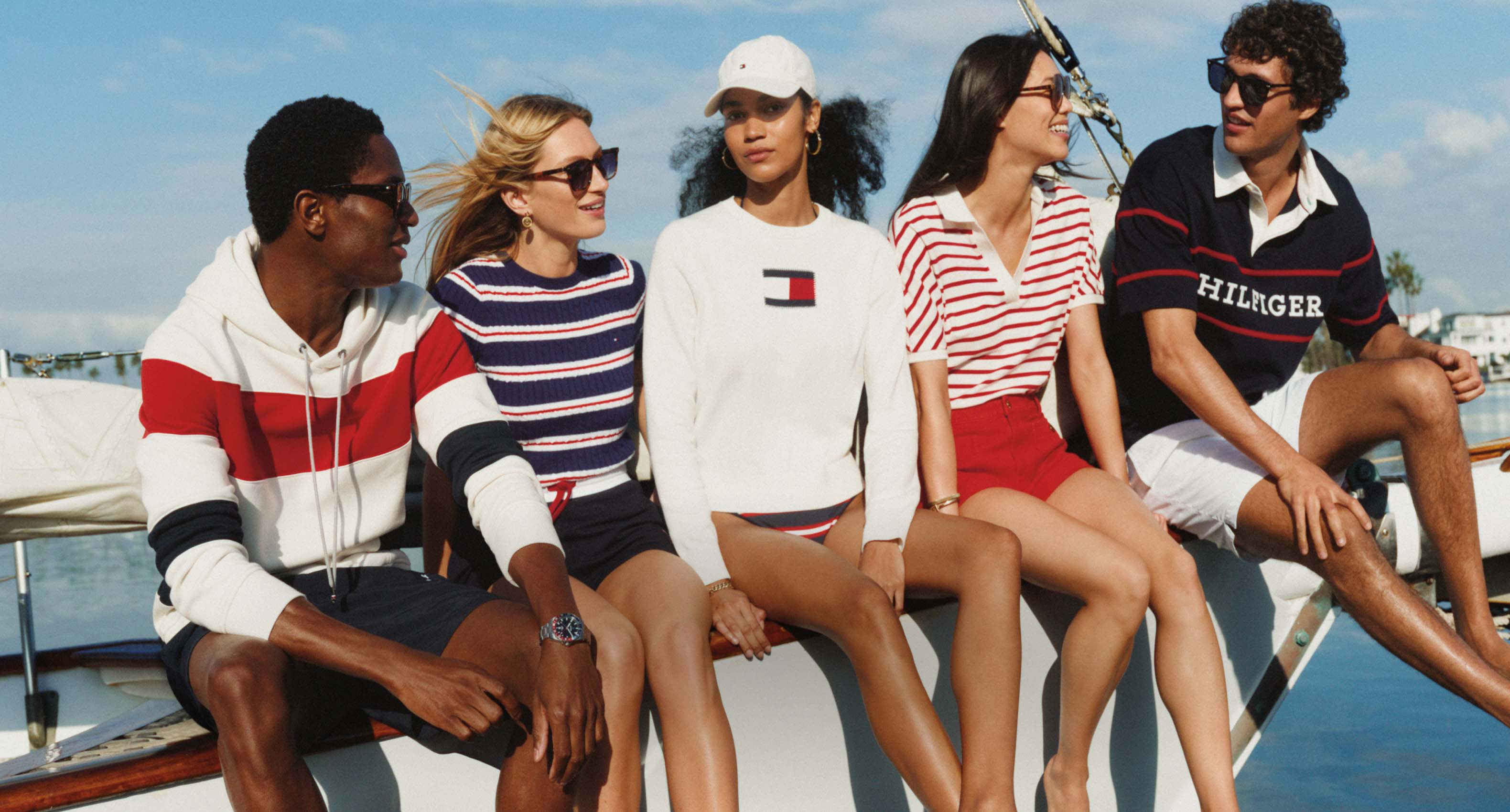 Five models sitting together on a sailboat wearing red, white, and navy Tommy Hilfiger summer styles against a sunny waterfront backdrop.
