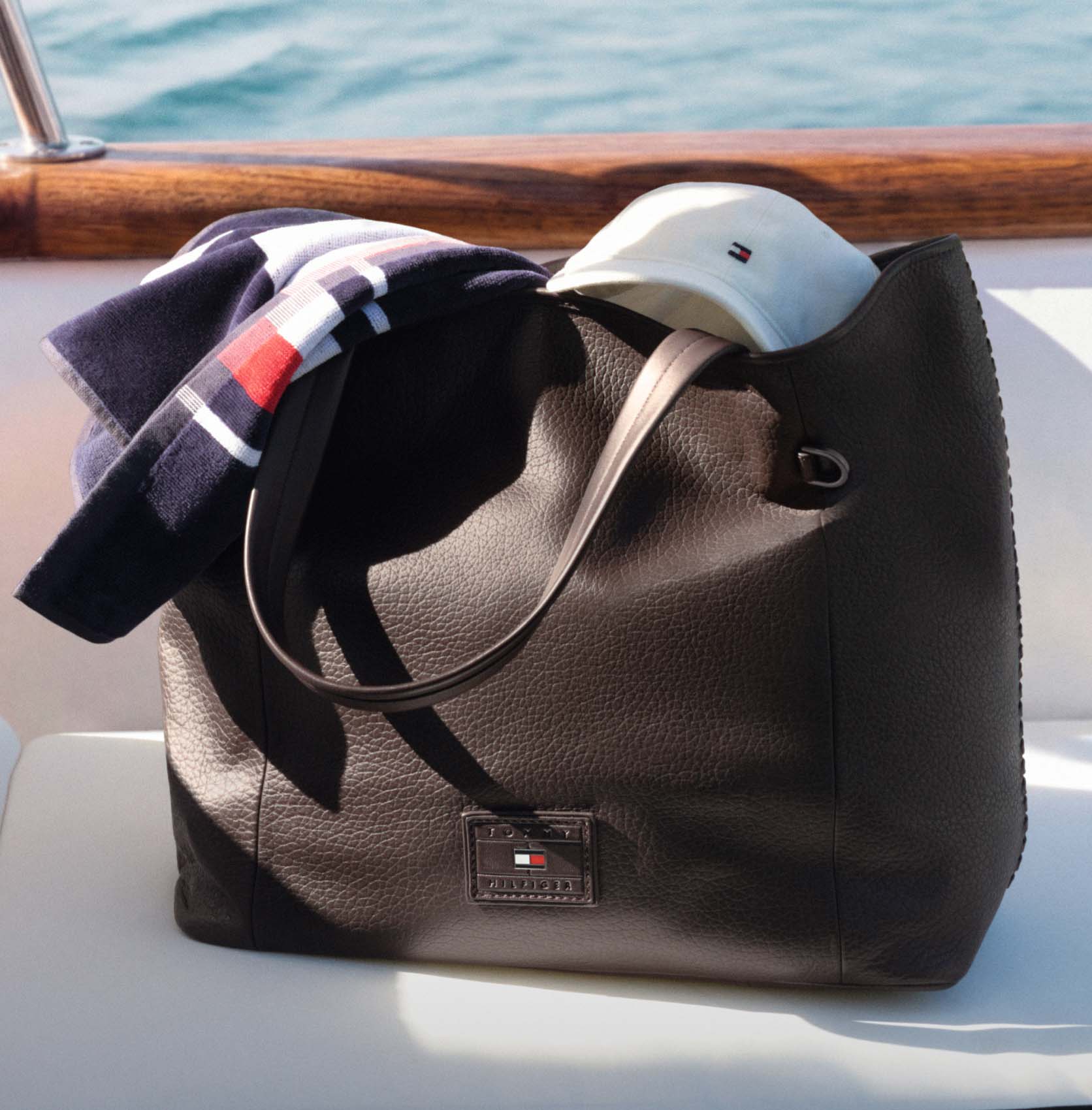 Tommy Hilfiger tote bag styled with folded apparel, scarves, and a baseball cap on a boat deck.