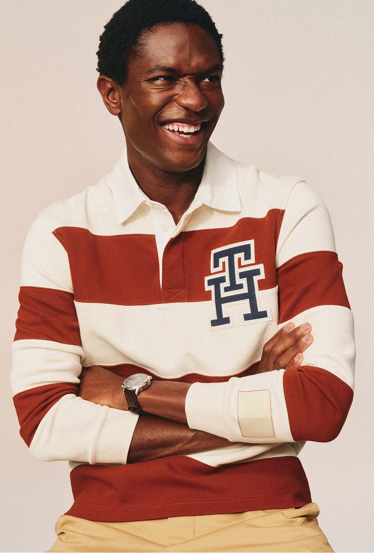 Male model smiling while wearing a cream rugby-style polo with red stripes and Tommy Hilfiger logo.
