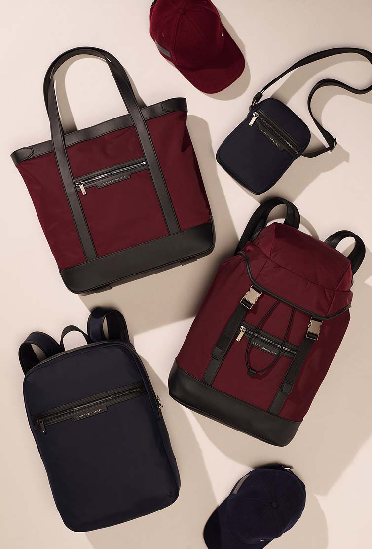 Assortment of Tommy Hilfiger tote bags, crossbody bags, and accessories arranged on a neutral background.