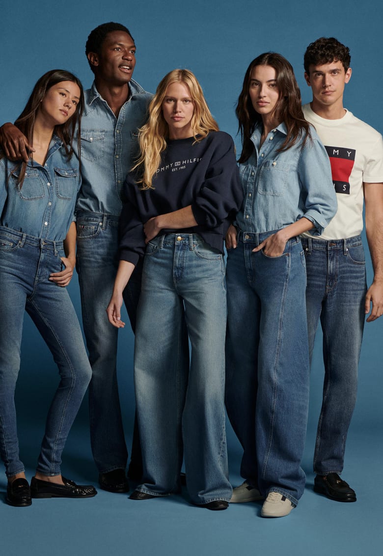 Group of five models wearing a variety of men's and women's Tommy Hilfiger denim jeans, shirts, and sweaters against a blue studio backdrop.