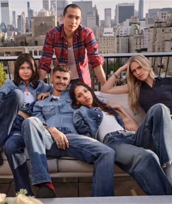 Five models wearing Tommy Hilfiger denim apparel pose together on a rooftop terrace with a city skyline in the background.