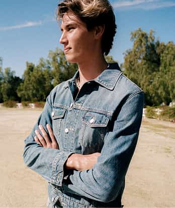 Man wearing a Tommy Jeans light blue denim shirt stands outdoors with a beach and blue sky in the background.