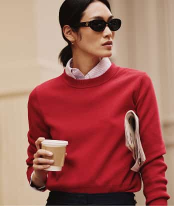 Woman wearing a Tommy Hilfiger red sweater, black trousers, and sunglasses holds a takeaway coffee outdoors.
