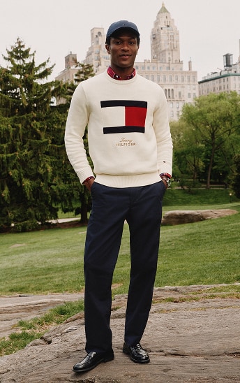 Man standing outdoors wearing a cream sweater with a large Tommy Hilfiger flag logo and navy pants.
