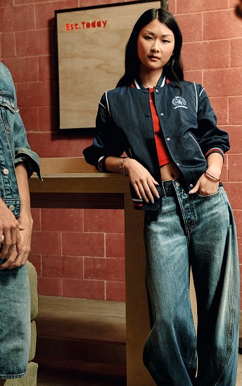Woman wearing a Tommy Jeans denim jacket and jeans while sitting outside against a brick wall.