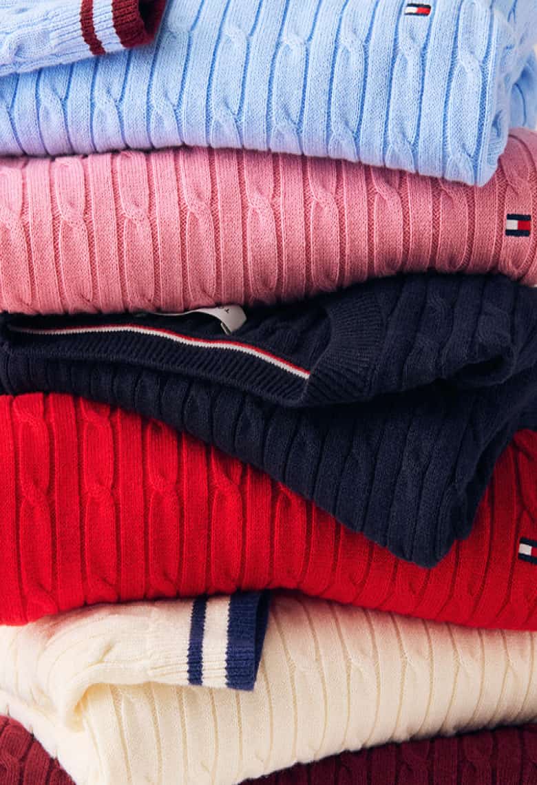 Two images side-by-side: Left side - Folded Tommy Hilfiger cable-knit sweaters in tan, purple, taupe, green, and blue stacked to showcase colors and texture. Right side - Smiling model wearing a Tommy Hilfiger red cable-knit sweater layered over a white collared shirt.