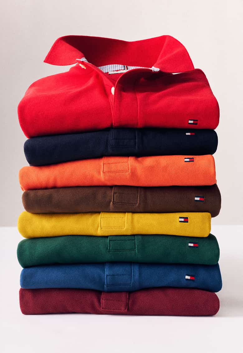 A side-by-side image collage: Left side - Smiling model wearing a Tommy Hilfiger dark green short-sleeve polo shirt with striped collar and sleeve trim against a neutral background. Right side - Stack of folded Tommy Hilfiger polo shirts in red, navy, orange, brown, yellow, green, blue, and burgundy with a red polo displayed on top.