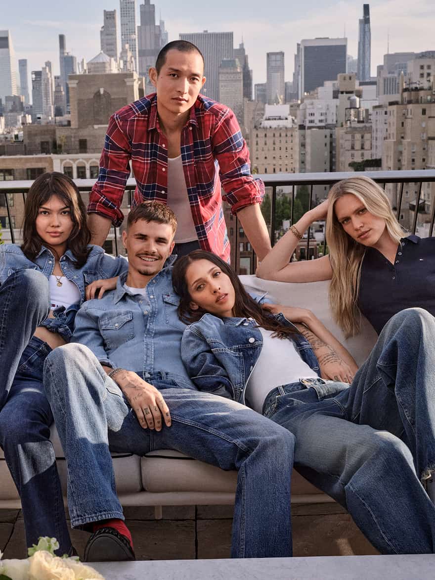 An image of Romeo Beckham and models wearing all denim Tommy Hilfiger outifts sitting on a couch outdoors on a rooftop, with a city skyline in the background.