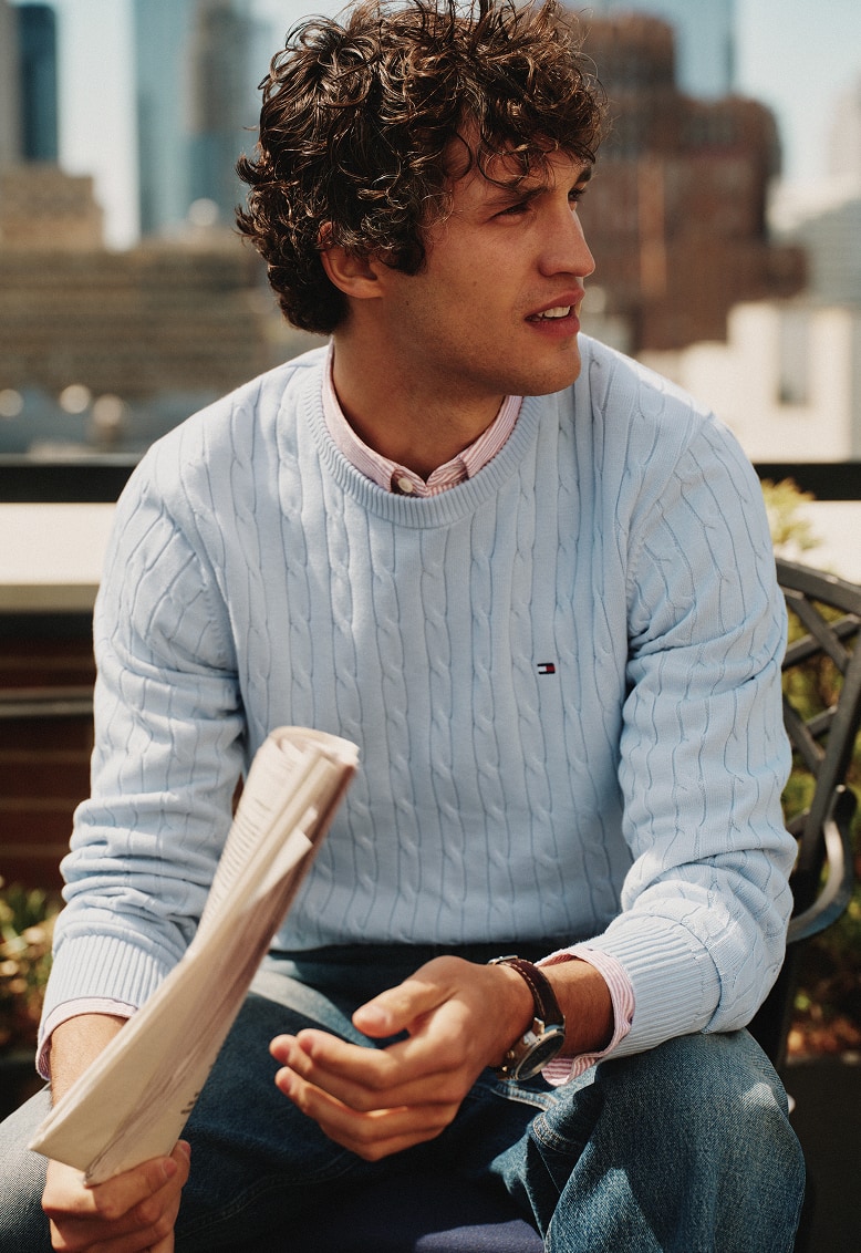 Model sitting outdoors wearing a Tommy Hilfiger light blue cable-knit sweater over a collared shirt while holding a newspaper.