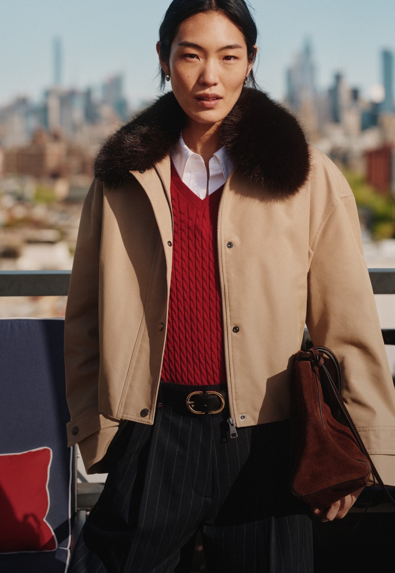 Model wearing a Tommy Hilfiger beige jacket with a dark faux-fur collar over a red cable-knit sweater and white shirt, standing on a rooftop with a city skyline in the background.