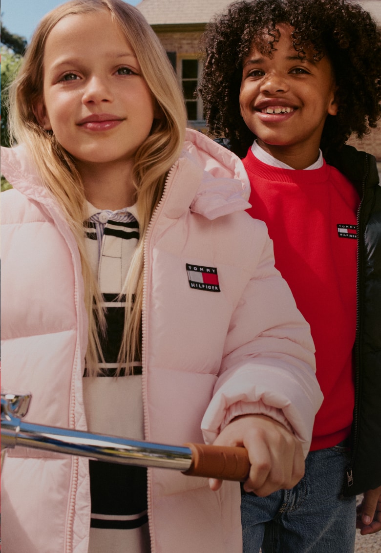 Two smiling children wearing Tommy Hilfiger outerwear stand outdoors beside a bicycle.