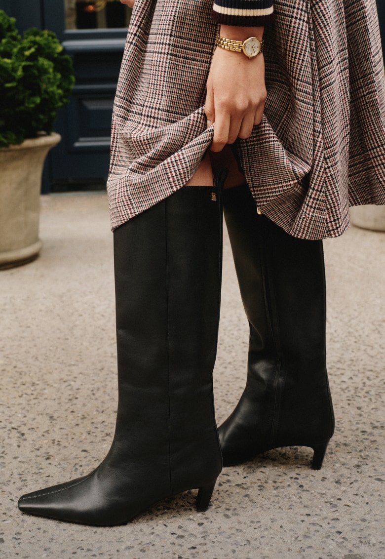 Close-up of a person wearing a Tommy Hilfiger black knee-high leather boots, holding a brown handbag, and wearing a plaid skirt.