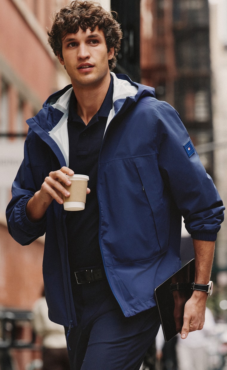 Man wearing a Tommy Hilfiger blue performance jacket carries a takeaway coffee while walking outdoors.