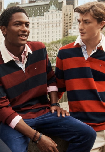 Two men sit on outdoor steps wearing Tommy Hilfiger striped rugby-style varsity polos in navy, red, and white.