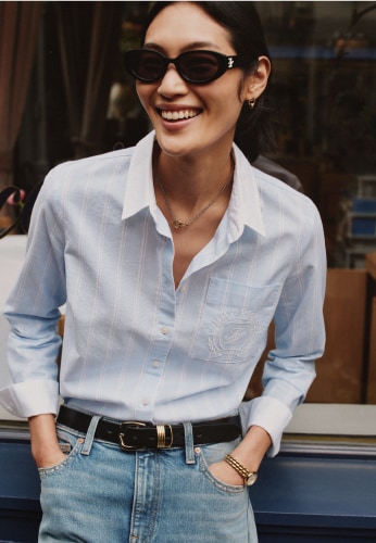 Woman wearing a Tommy Hilfiger light blue button-down shirt, jeans, and sunglasses smiles while walking outdoors.