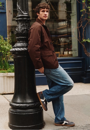 Man in a Tommy Hilfiger brown jacket, jeans, and sneakers leans against a lamppost on a city street.