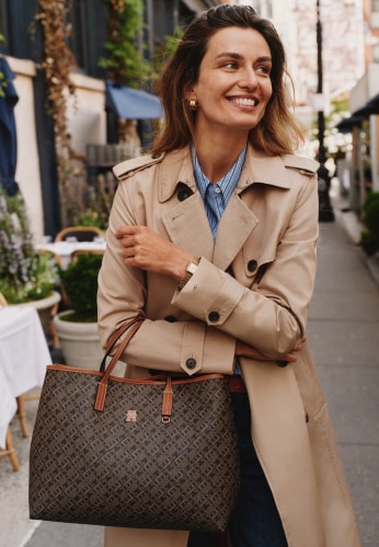 Woman wearing a Tommy Hilfiger beige trench coat carries a brown monogram tote bag while walking through the city.